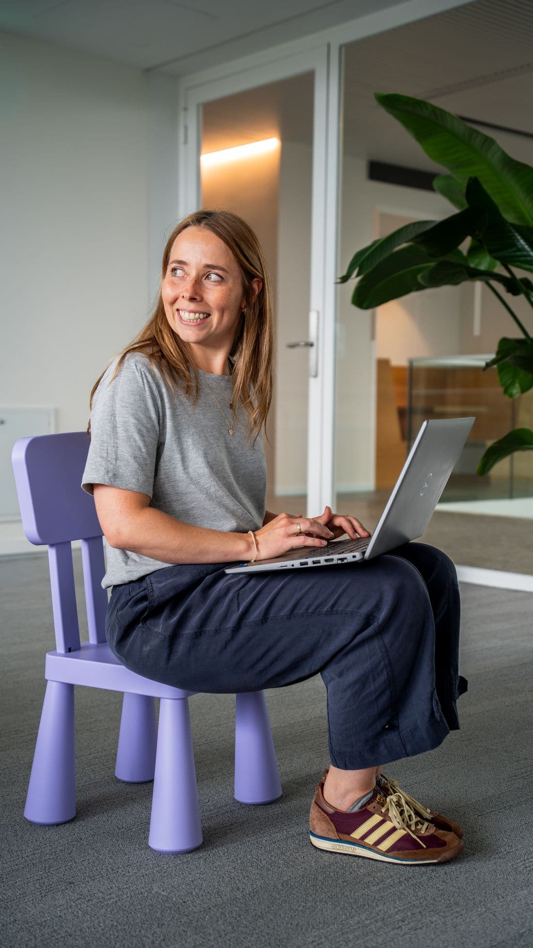Vrouw met laptop op een paars kinderstoeltje, lachend over haar schouder