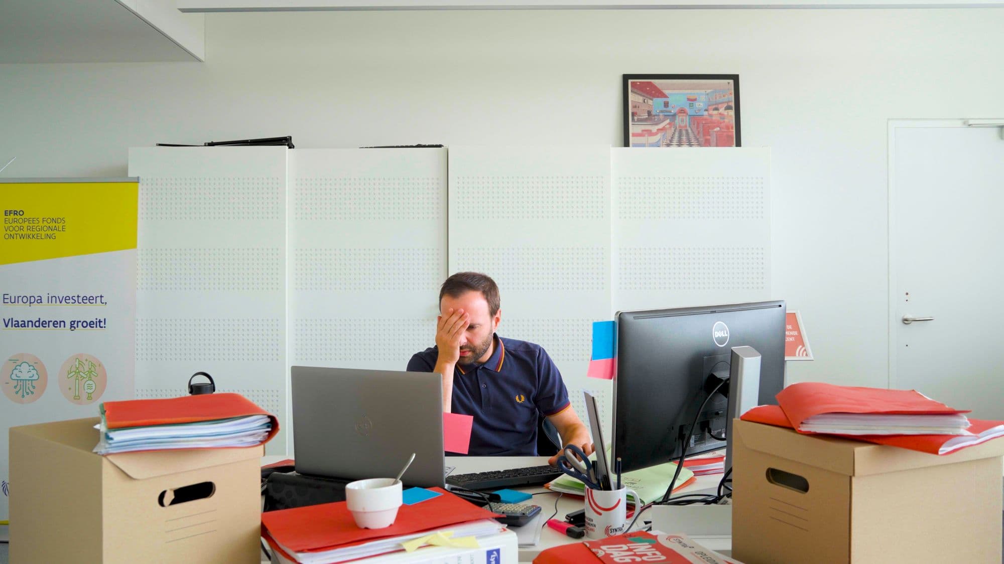 Medewerker met de hand voor zijn gezicht achter een bureau vol mappen en dozen